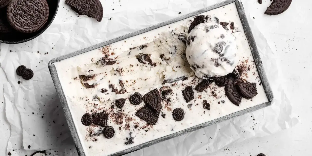 Cremiges Oreo Eis in einer Metallform, garniert mit ganzen und zerbrochenen Keksen. Ein Löffel Eis liegt obenauf.