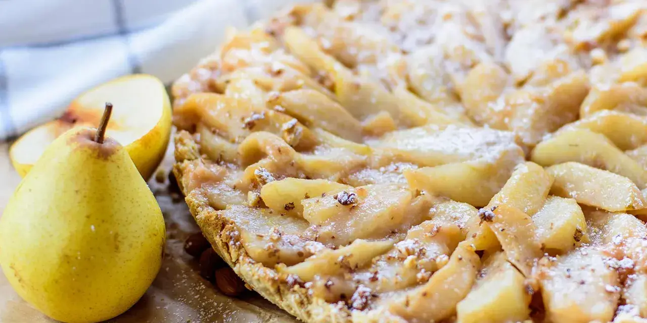 Délicieuse tarte aux poires, saupoudrée de sucre glace, avec des amandes et des poires fraîches à côté.