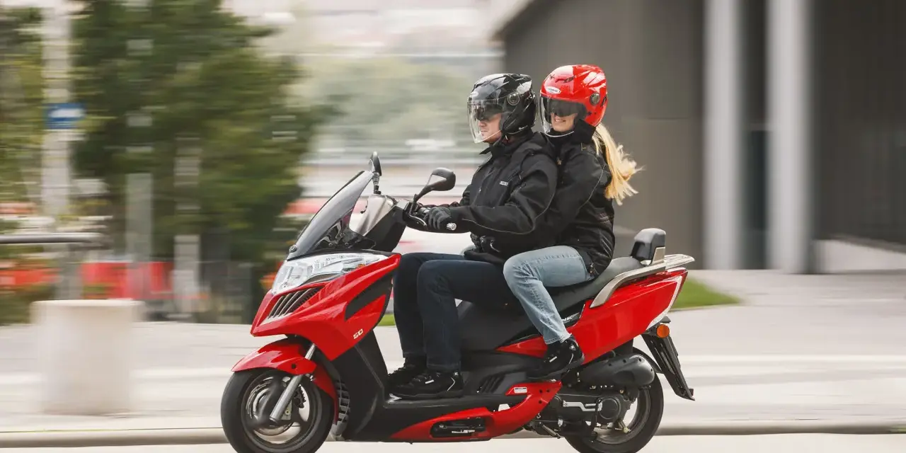Para jadąca na czerwonym skuterze 125, ubrana w kurtki i kaski, cieszy się jazdą.