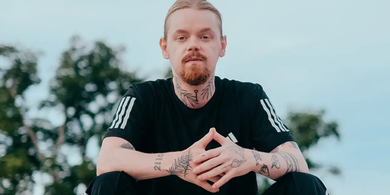 Mężczyzna z rudą brodą i blond włosami, ubrany w czarną koszulkę z trzema paskami. Na szyi i rękach widoczne tatuaże, w tym klucz i napis "kuqe 2115".
