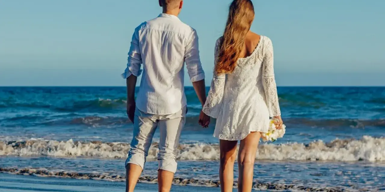 Para idąca plażą, trzymająca się za ręce. Ich miłość grecka kwitnie na tle błękitnego morza i nieba.