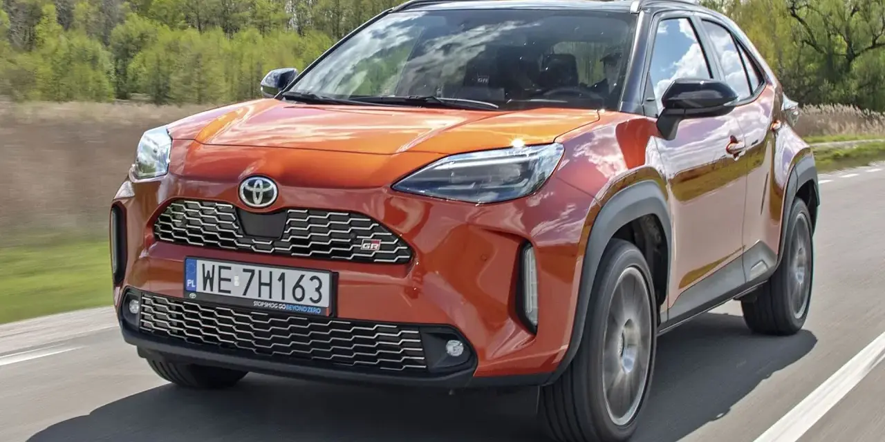Pomarańczowa Toyota Yaris Hybrid GR Sport pędzi po drodze, otoczona zielenią.