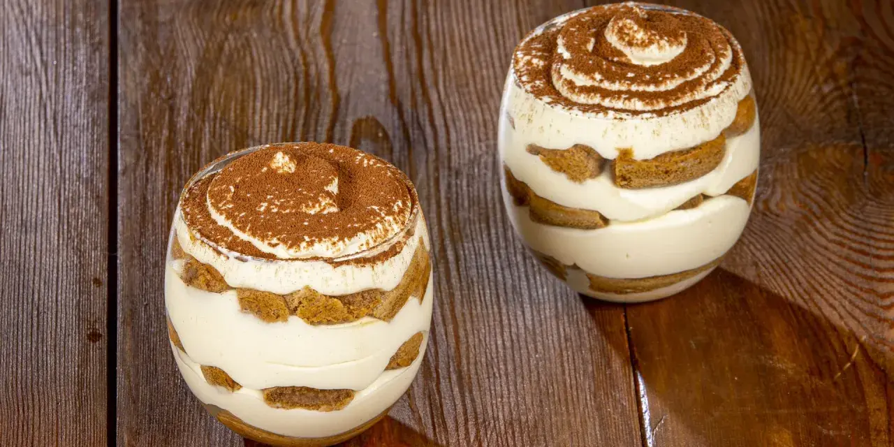 Due bicchieri di tiramisù senza uova, stratificati con crema e savoiardi, spolverati di cacao su un tavolo di legno.