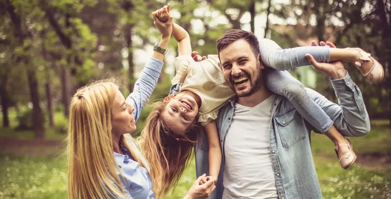 Glückliche Familie im Park. Vater trägt Tochter auf den Schultern, Mutter hält ihre Hände. Kein Anzeichen von Einzelkind-Syndrom, nur pure Freude.