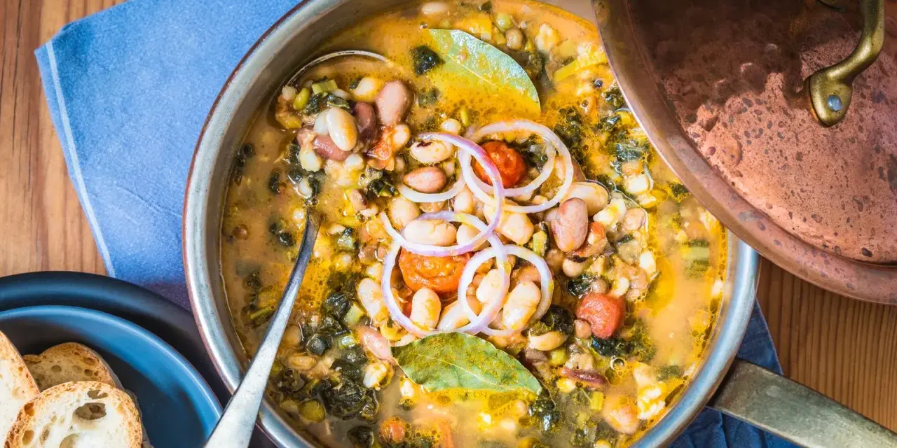 Zuppa ricca con fagioli, verdure e spezie, servita in una pentola di rame. Un piatto confortante di acqua cotta, guarnito con cipolla rossa e alloro.
