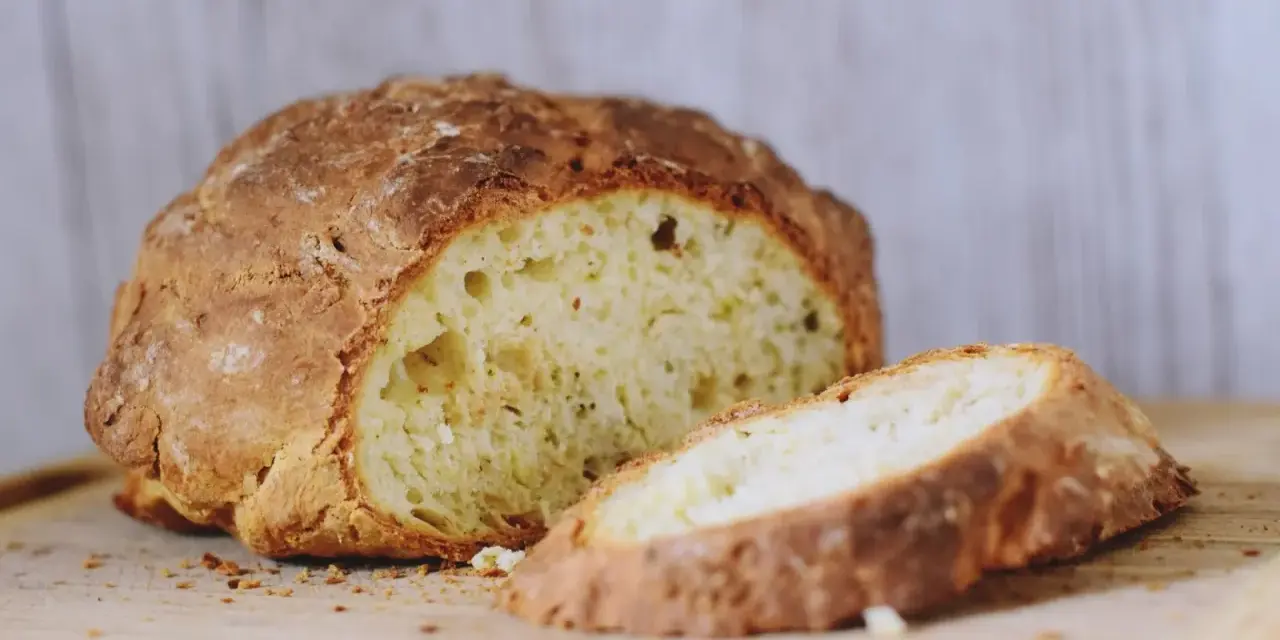 Chrupiący, złocisty bochenek soda bread, z jednym plastrem odciętym, na drewnianej desce.