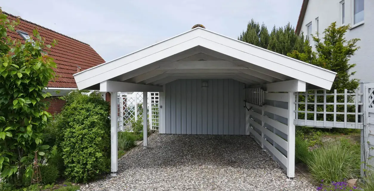 A white wooden carport with a gravel driveway, surrounded by greenery and a white lattice fence. This image shows a finished carport, a great example of how to build a carport.