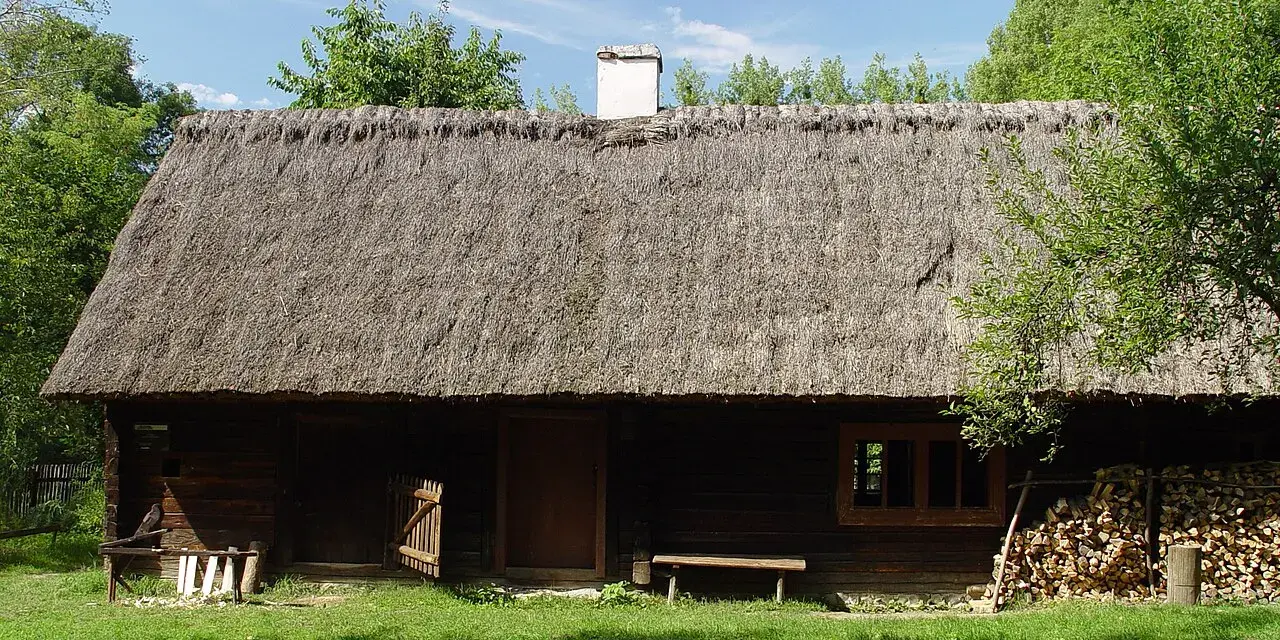 Drewniana chata z krytym strzechą dachem, część ekspozycji Muzeum Wsi Opolskiej. Obok stos drewna i ławka.