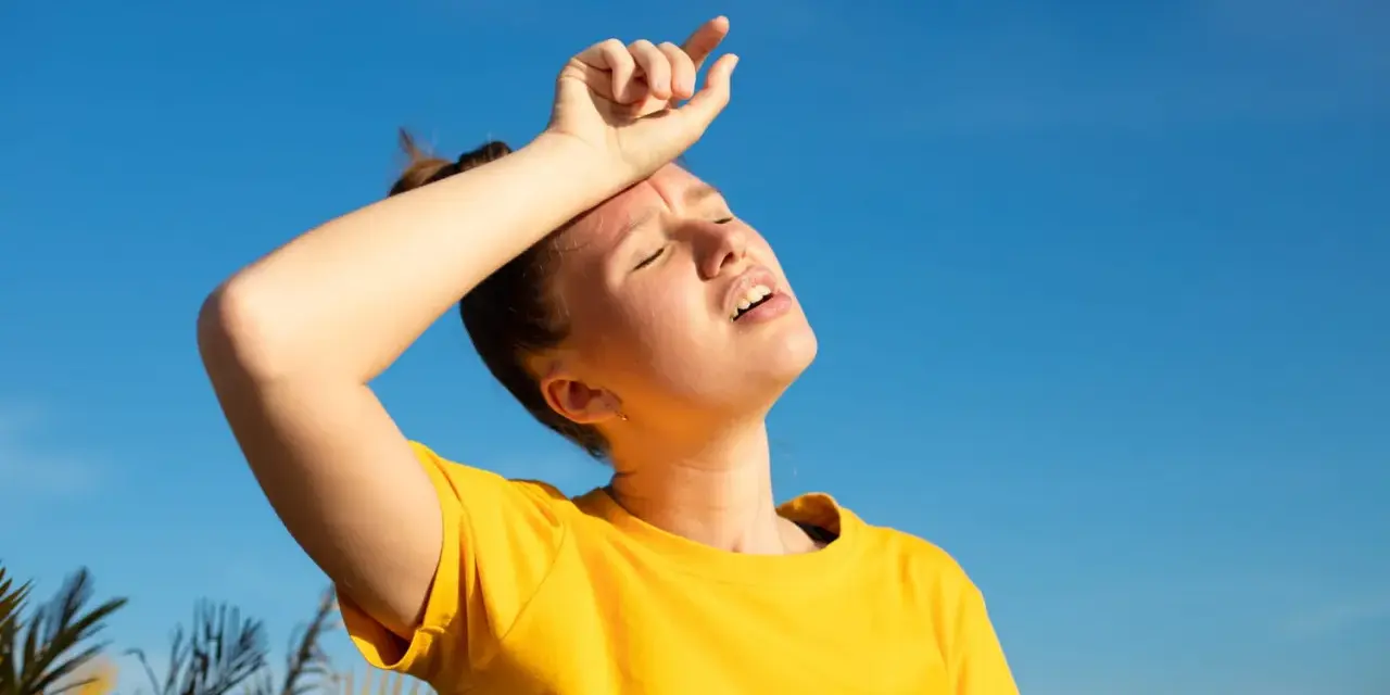 Une jeune femme en t-shirt jaune, le visage marqué par la chaleur, essuie son front sous un ciel bleu intense. La durée d'une insolation est palpable.