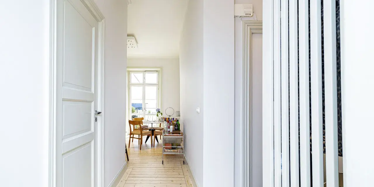 Ljus hall med trägolv och vita dörrar, perfekt för en renovera hall. Genom en valvöppning syns ett runt bord med stolar och ett fönster.