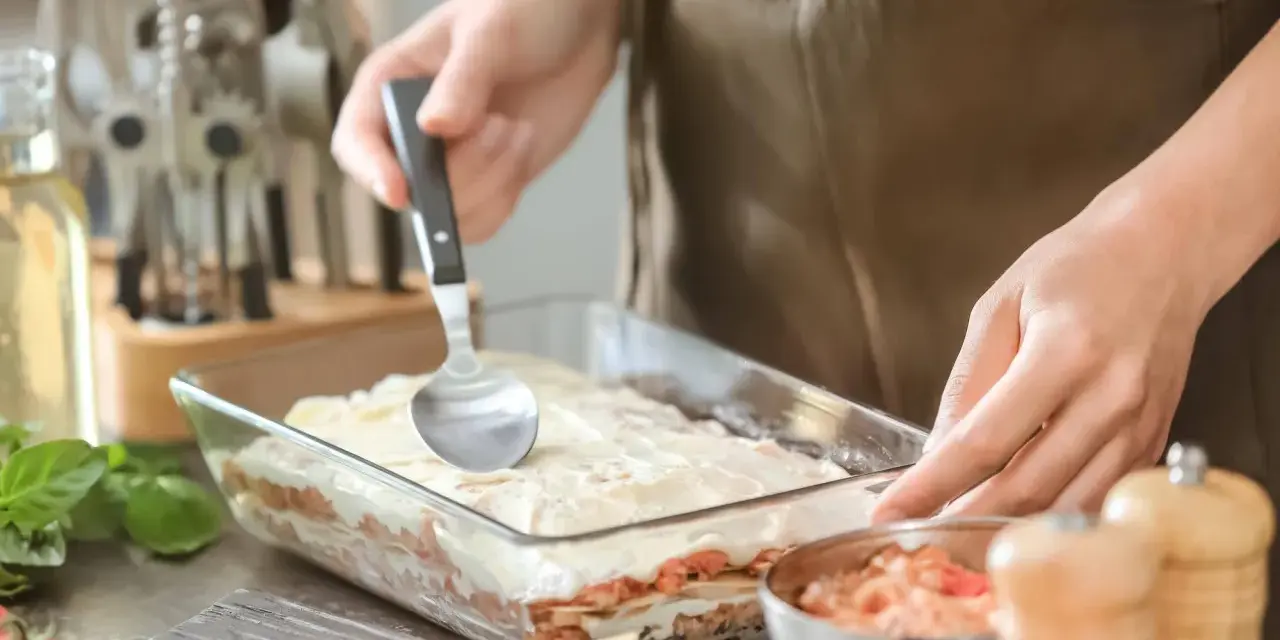Osoba nakłada sos na warstwy makaronu i mięsa, przygotowując do pieca lasagne.