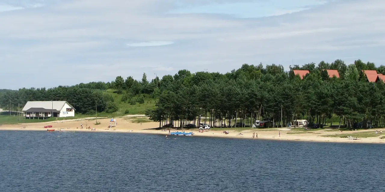 Plaża nad jeziorem Chańcza tętni życiem. Ludzie odpoczywają na piasku, kąpią się w wodzie, a w tle widać domki i las.