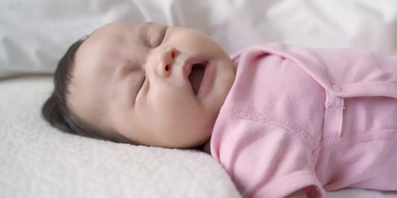Ein süßes neugeborenes Baby in rosa Strampler niest. Die Augen sind geschlossen, der Mund leicht geöffnet.