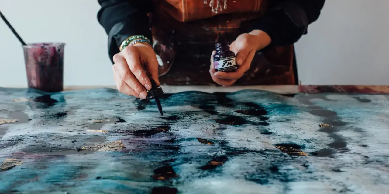 Une artiste crée une **aquarelle sur toile** aux teintes bleues et dorées, ajoutant des touches d'encre avec un compte-gouttes.