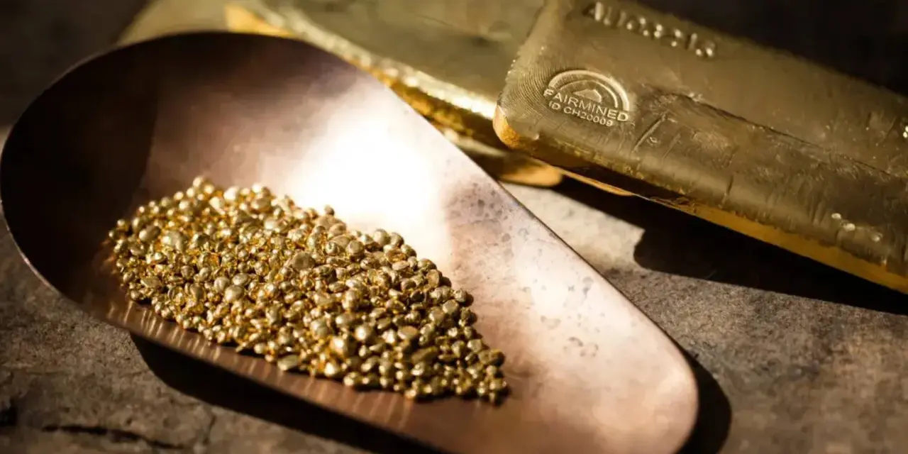 Piccoli granelli scintillanti di oro riciclato riposano in una paletta di rame, accanto a lingotti dorati.
