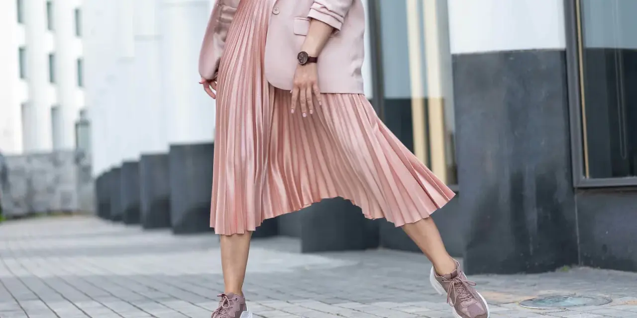 Kobieca stylizacja: różowa plisowana spódnica, marynarka i sportowe buty. Idealne połączenie elegancji i wygody, pokazujące, jakie buty do plisowanej spódnicy pasują najlepiej.