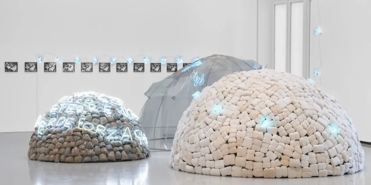 Installation d'art povera : dômes en pierres et en tissu, ornés de néons bleus, sous une structure transparente.