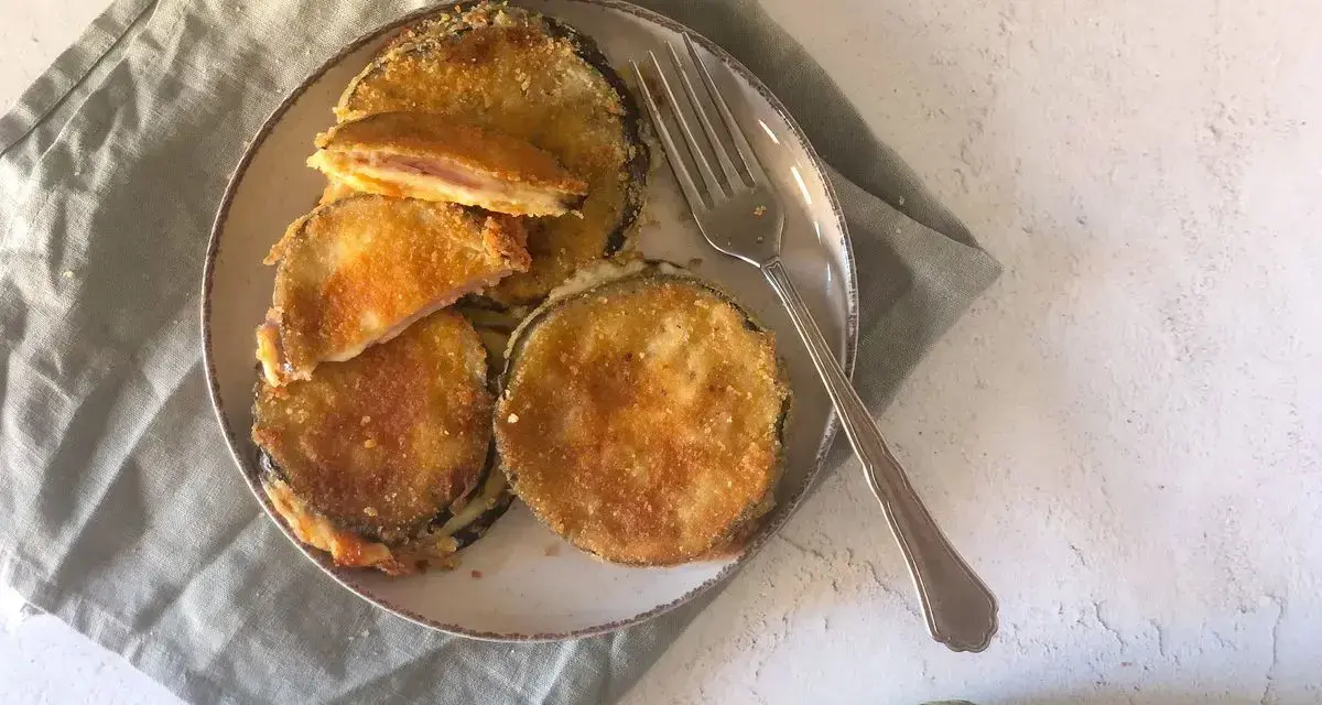 Cordon bleu di melanzane impanati e fritti, ripieni di prosciutto e formaggio filante, serviti su un piatto con una forchetta.