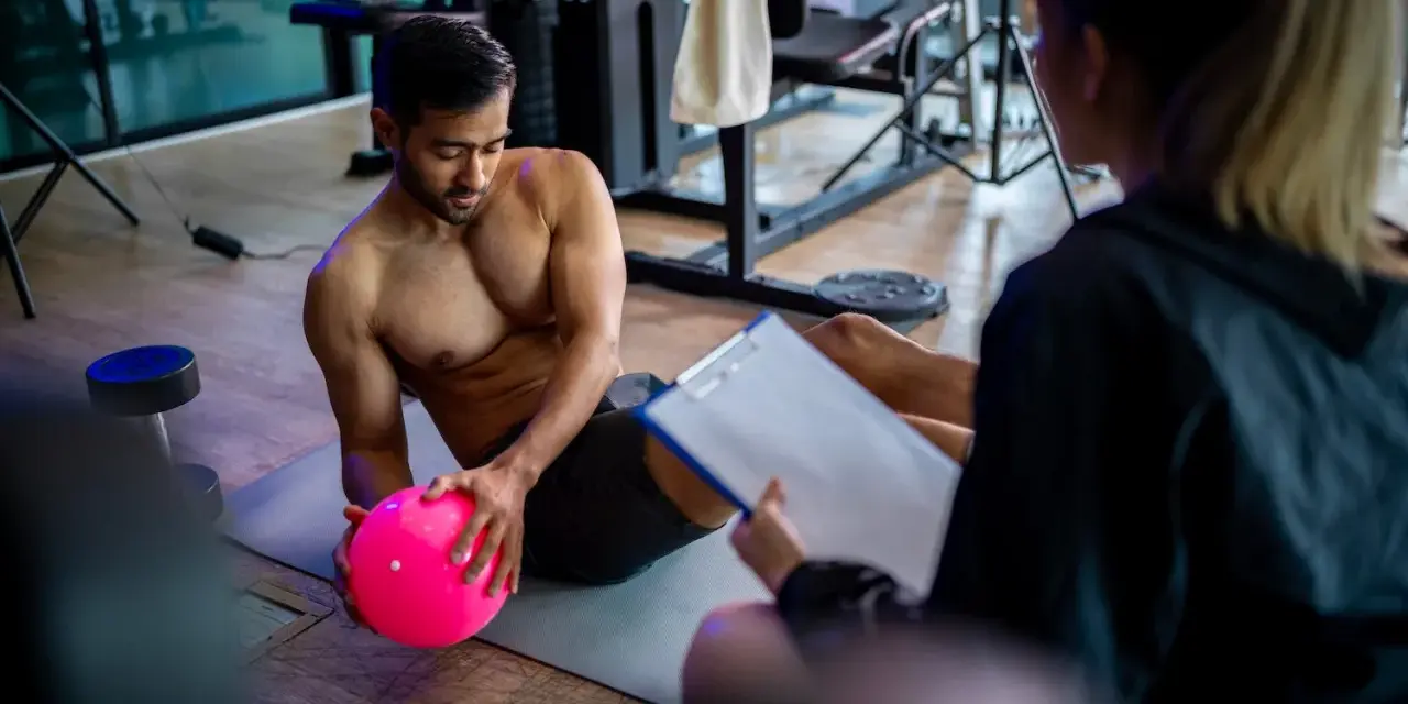 Man med rosa boll på gymmatta, tränare med clipboard. De följer sitt gymprogram.