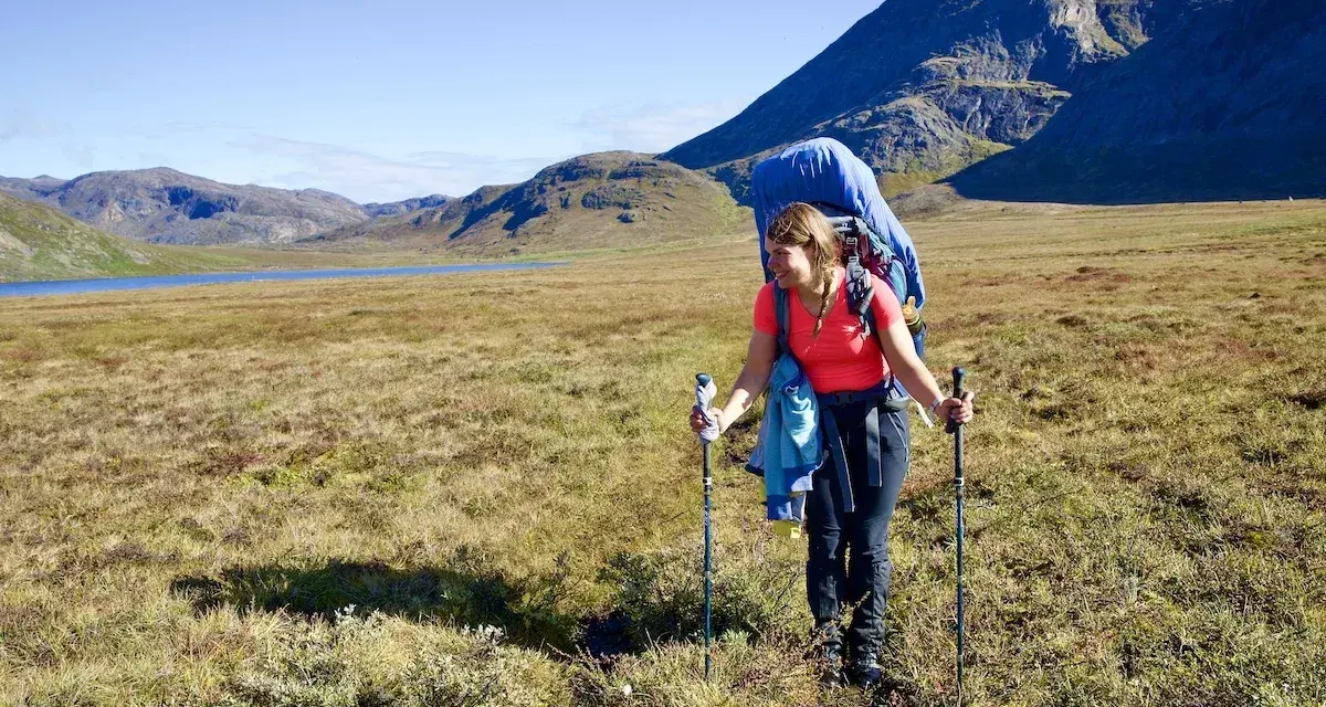 Frau wandert mit großem Rucksack und Stöcken über einen grasbewachsenen Hügel. Im Hintergrund ein See und Berge. Ein Abenteuer auf dem Arctic Circle Trail.