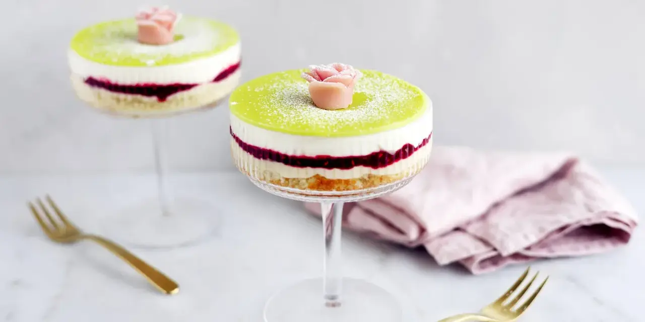 Två vackra prinsesstårtor i glas, med lager av sockerkaka, sylt och grädde, toppade med grön marsipan och en marsipanros.
