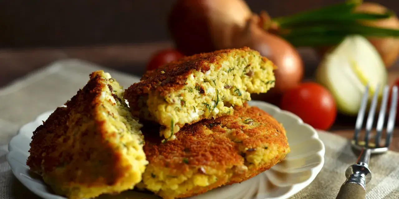 Pyszny kotlet jajeczny z ziołami, podany na talerzyku. W tle cebula i pomidorki.
