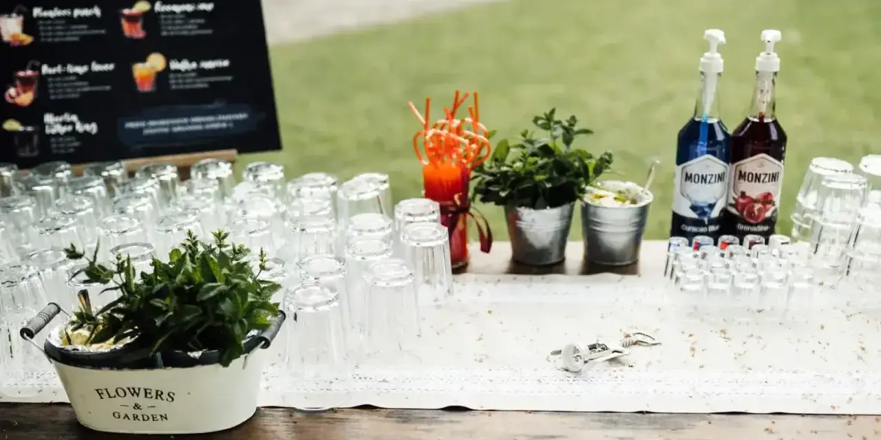 Stół z kieliszkami, butelkami syropów i miętą, gotowy na przygotowanie drinków na wesele. Przepisy na drinki są widoczne na tablicy w tle.