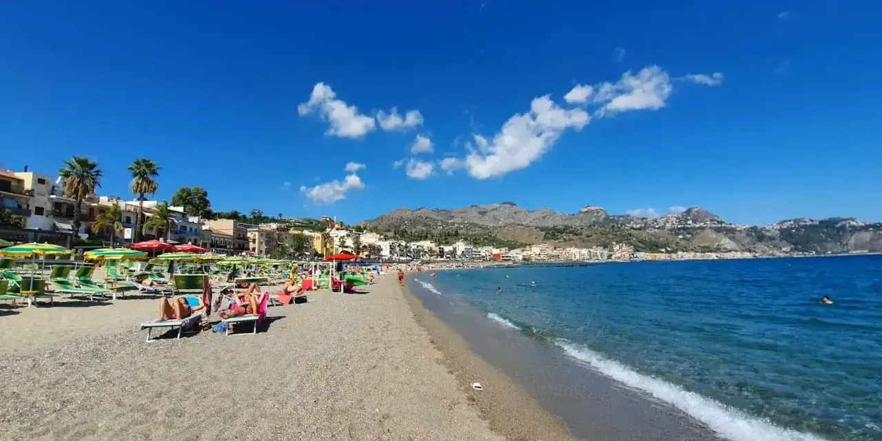 Spiaggia affollata a Giardini di Naxos, con lettini, ombrelloni e persone che si rilassano sotto il sole siciliano.