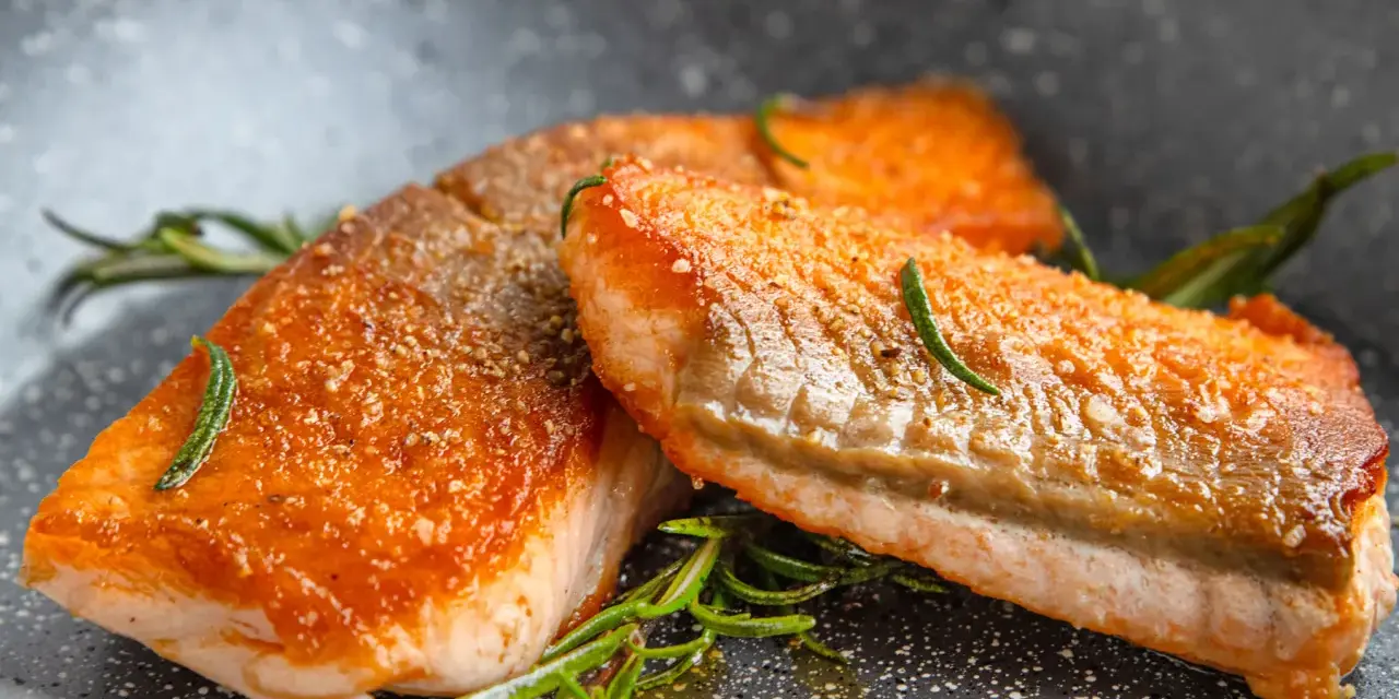 Zwei saftige Lachsfilets mit knuspriger Haut, perfekt gebraten, liegen in einer Pfanne mit Rosmarin und Öl.