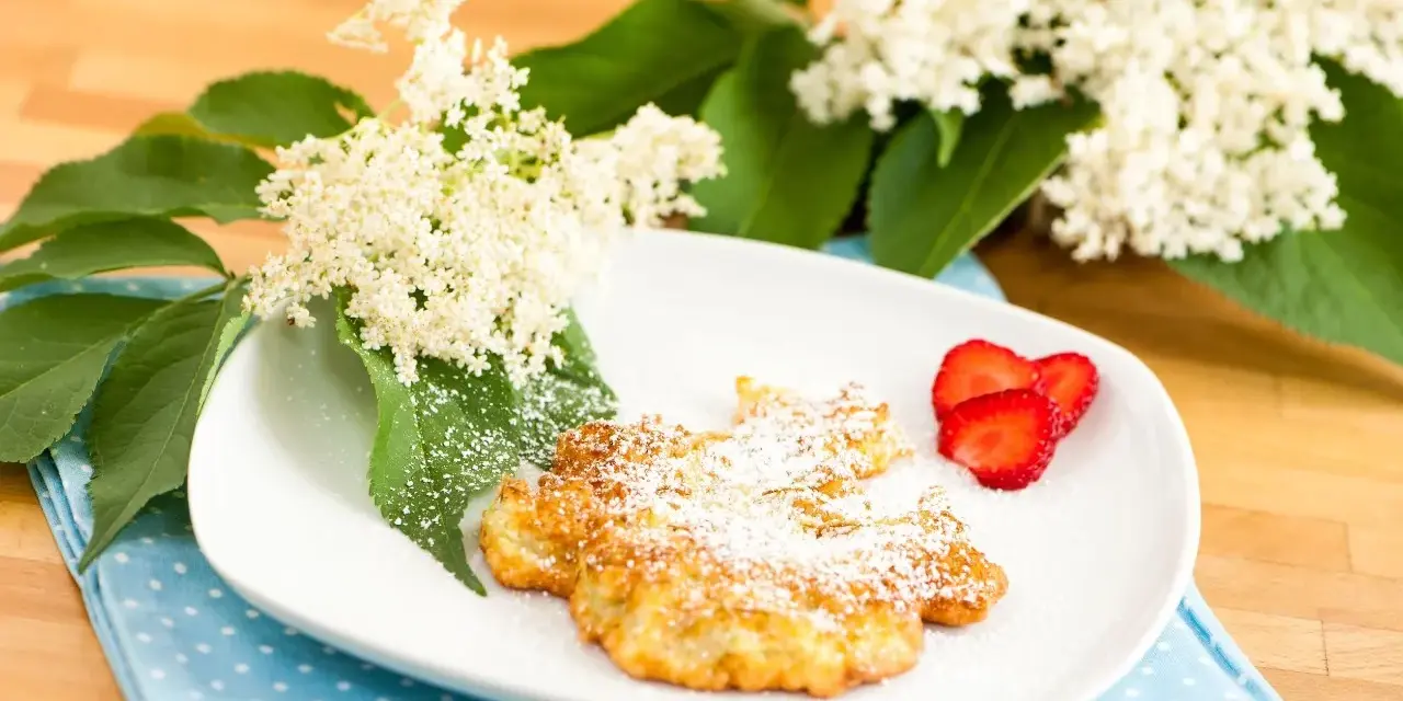 Fiori di sambuco fritti, spolverati di zucchero a velo, serviti su un piatto bianco con fragole fresche e decorati con fiori di sambuco.