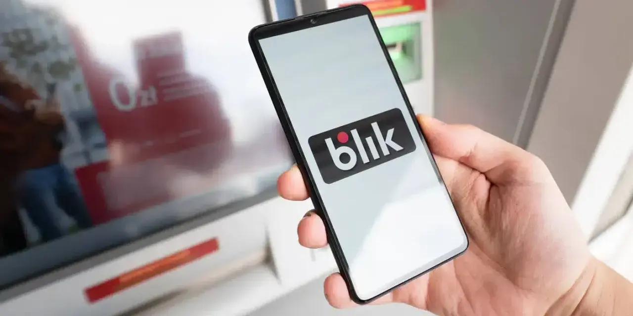 Dłoń trzyma smartfon z logo BLIK przed bankomatem.