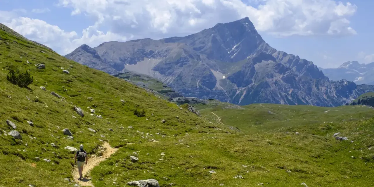 Escursionista su sentiero erboso, con imponente gruppo montuoso delle Dolomiti sullo sfondo.