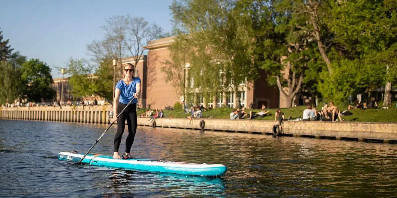 Frau genießt Stand Up Paddling auf einem Fluss, während Menschen am Ufer entspannen.