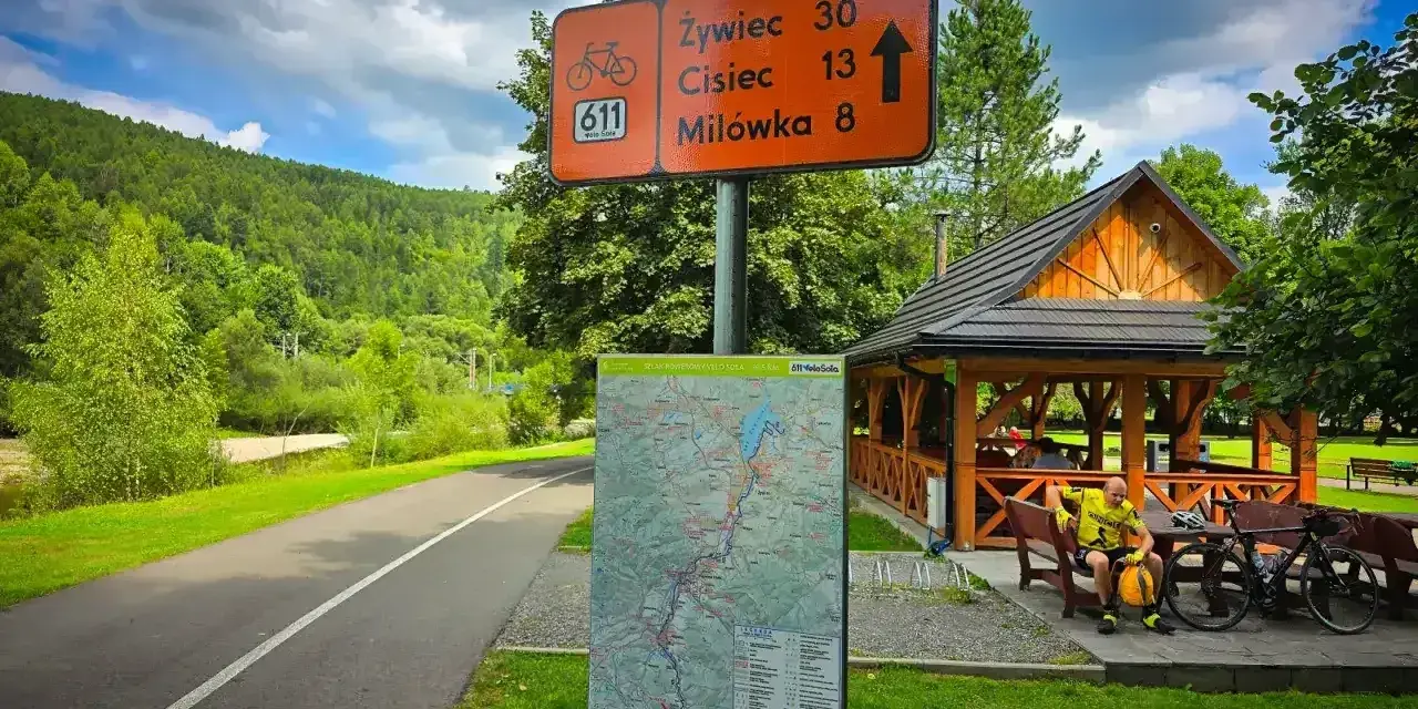 Znak szlaku rowerowego 611 Velo Soła wskazuje kierunki. Obok mapa i wiata z rowerzystą.