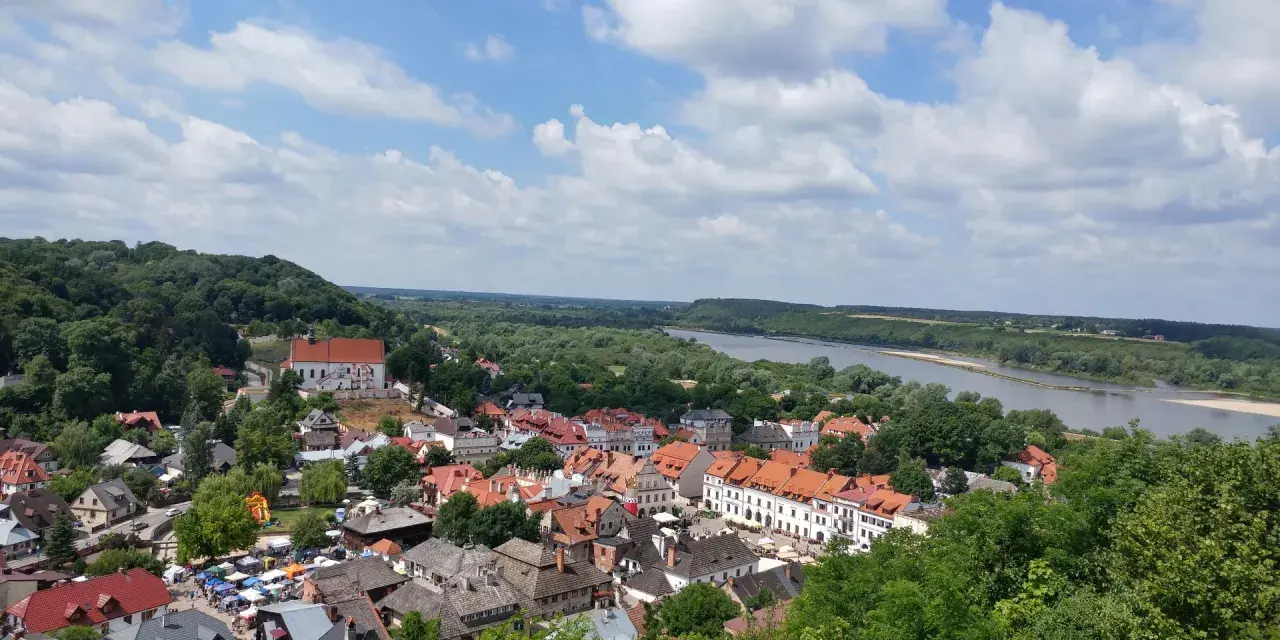 Widok na Kazimierz Dolny z górami Trzech Krzyży w tle. Rzeka Wisła, zielone wzgórza i urokliwe miasteczko z czerwonymi dachami.