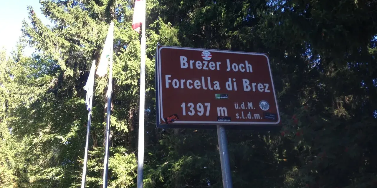 Cartello indicante "Forcella di Brez" a 1397 m, con bandiere e alberi sullo sfondo.