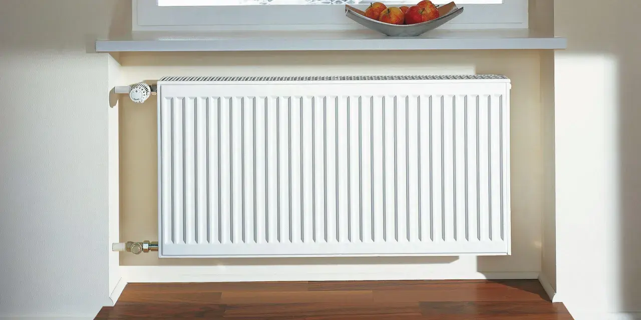 Radiador blanco de aerotermia con aletas de aluminio, bajo una ventana con persiana y un cuenco de manzanas.