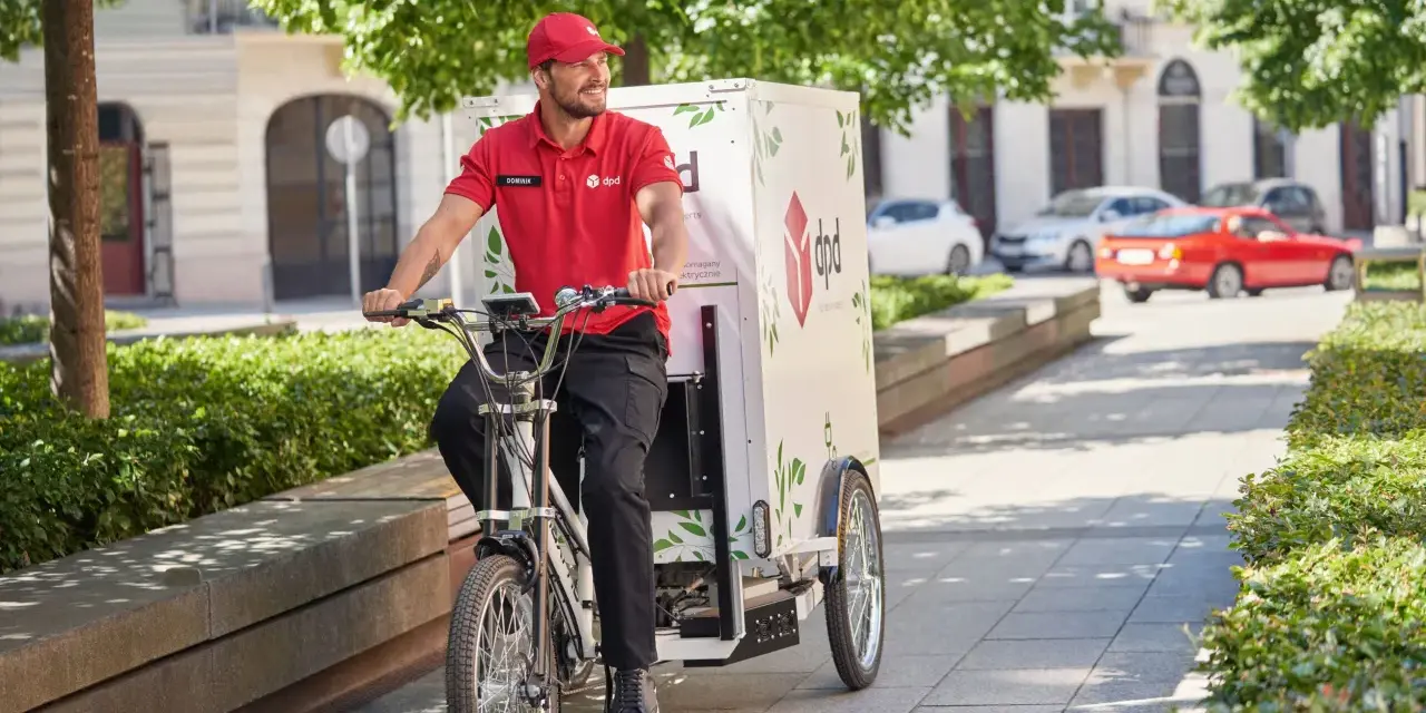 Uśmiechnięty kurier rowerowy DPD w czerwonej koszulce jedzie ulicą z zielonymi drzewami.