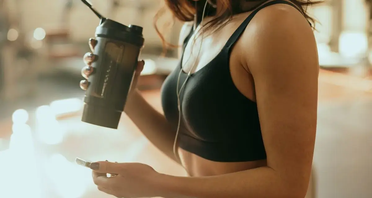 Kobieta w sportowym topie trzyma shaker, zastanawiając się, kiedy pić białko przy odchudzaniu. Słucha muzyki na telefonie.