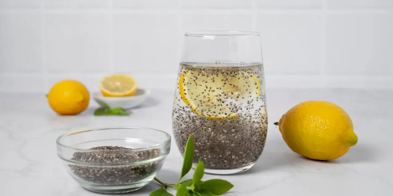 Vaso con agua de chia, rodaja de limón y semillas de chía en un bol. Limones y hojas verdes completan la escena.