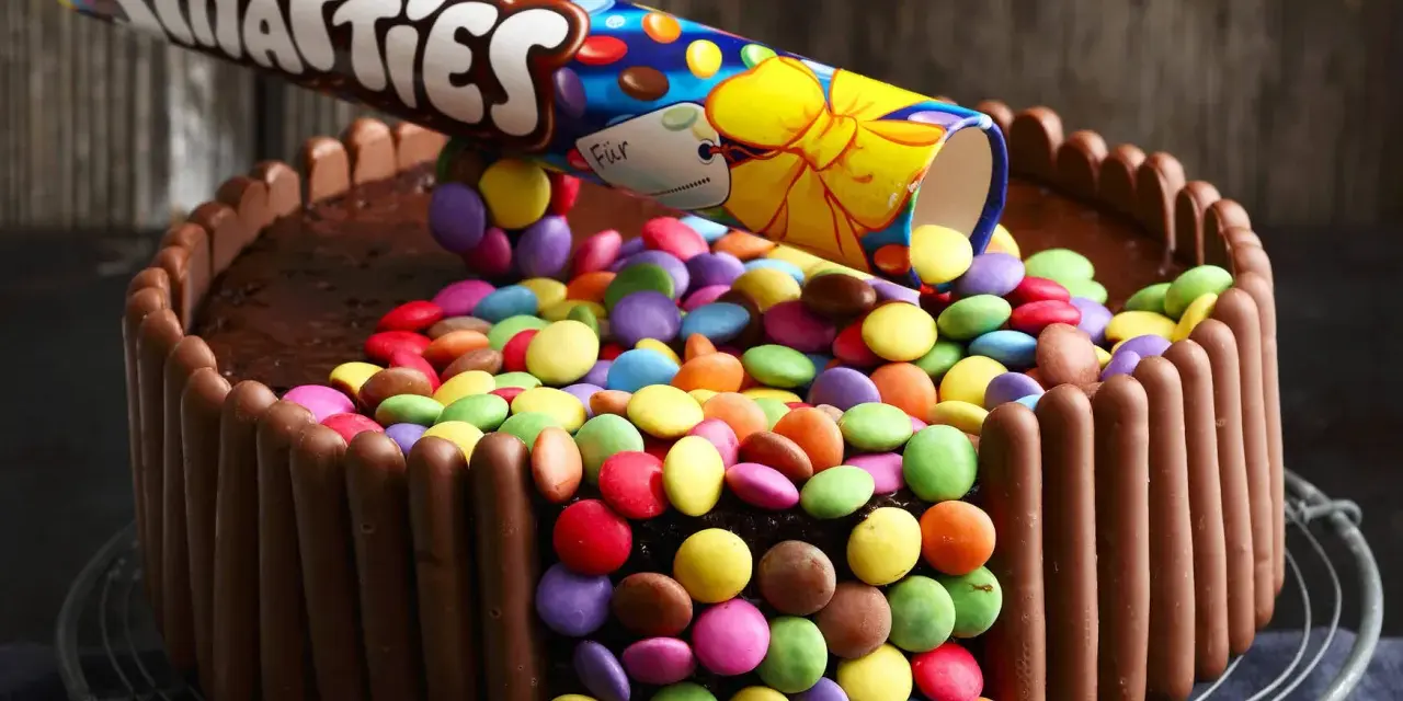 Eine einfache Geburtstagstorte für Jungs: Schokoladenkuchen, umhüllt von Schokoriegeln und bedeckt mit bunten Smarties, die aus einer aufgeschnittenen Rolle rieseln.