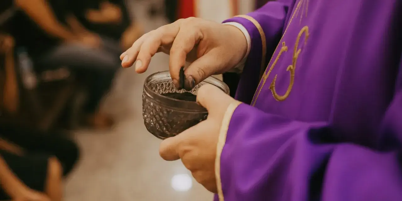 A priest in a purple robe takes ashes from a small bowl, preparing for the first day of Lent prayer.