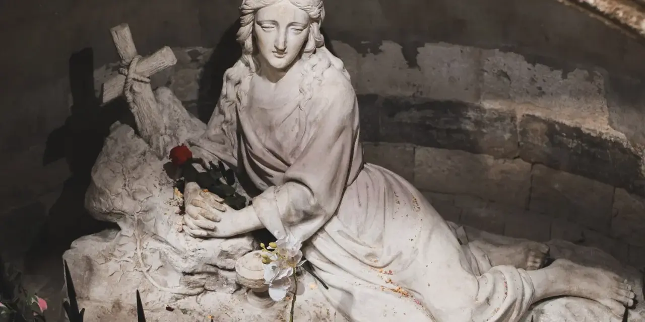 A stone sculpture depicts the serene body of Mary Magdalene, resting with a cross and a single red rose.