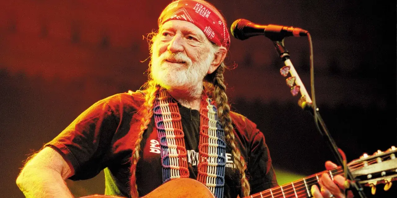 Willie Nelson mit roter Bandana und Zöpfen spielt Gitarre. Ein Mikrofon steht bereit für den Titel von Willie Nelson.