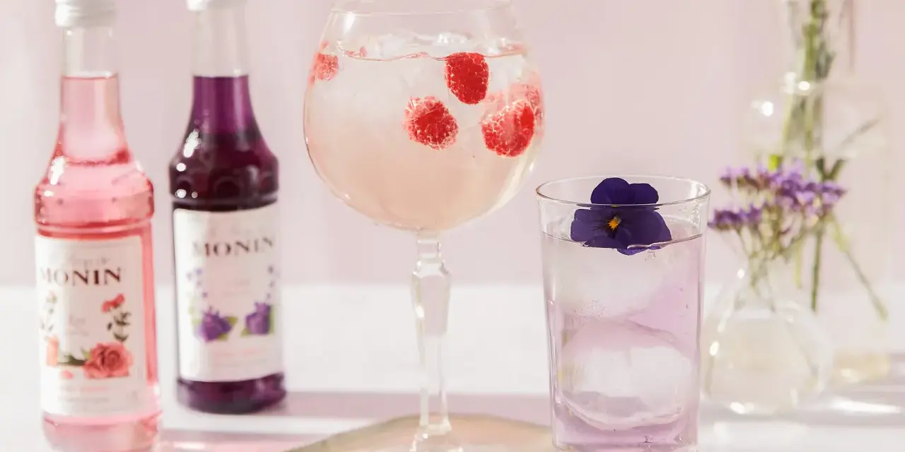 Dobre drinki przepisy: różowy i fioletowy syrop Monin, drink z malinami w kielichu, drink z fiołkiem w szklance, kwiaty w wazonach.