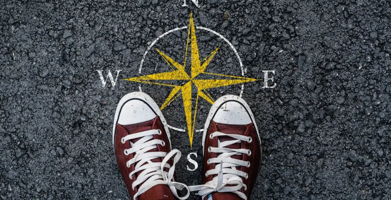 Feet stand on a compass, symbolizing the start of a journey. This image can represent the **mission creep meaning**, where initial goals expand unexpectedly.