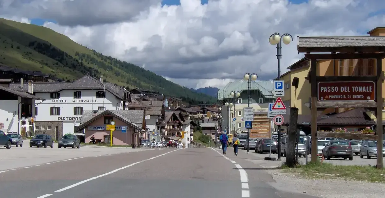 Strada verso il Passo del Tonale in moto, con case alpine e montagne sullo sfondo.