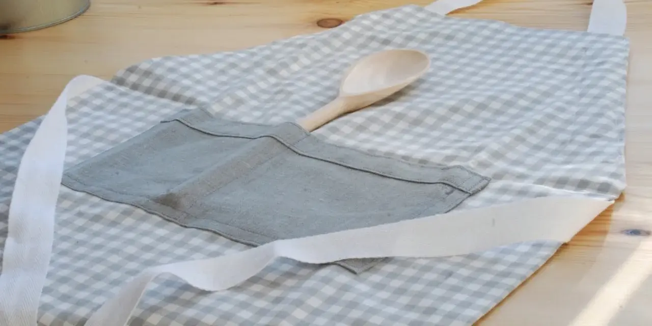 Gotowy fartuch kuchenny w kratkę, z drewnianą łyżką w kieszeni. Idealny poradnik, jak uszyć fartuch kuchenny.