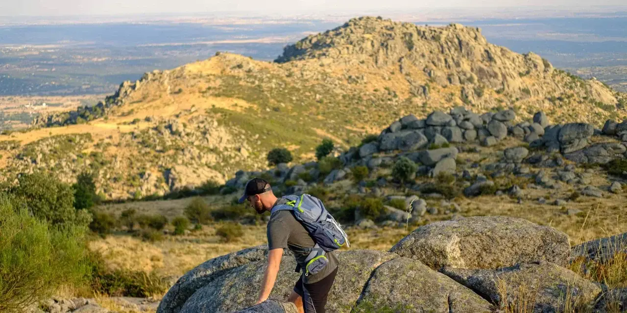 Un excursionista con mochila asciende por un terreno rocoso en la ruta Las Machotas, con un paisaje montañoso y un valle al fondo.
