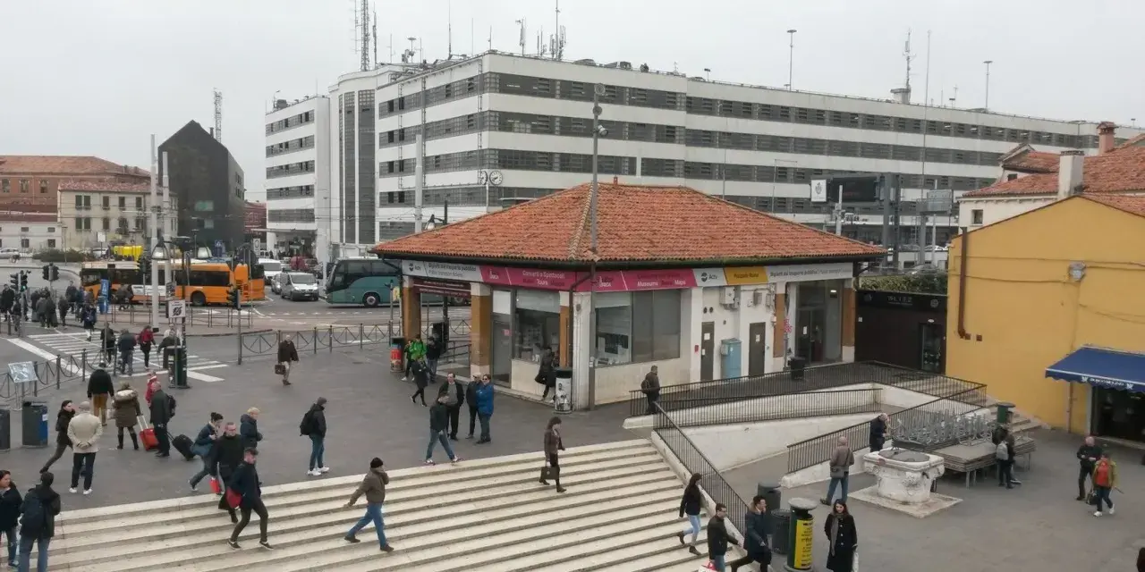 Persone attraversano una piazza a Venezia, con autobus e un edificio moderno sullo sfondo.